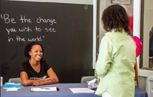 Woman behind a desk with a woman in front. "Be the change you want to see in the world" is written on a black board.