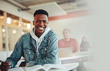 Young adult student sitting in a classroom and smiling with a pen in hand and open book.